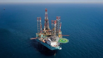 Offshore jack-up drilling rig with red and white derrick and green helipad surrounded by calm blue open sea