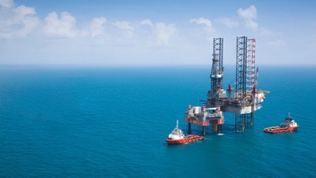 Aerial view of an offshore jack-up drilling platform with two red support vessels on a calm turquoise sea