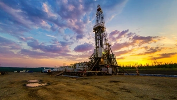 Oil and gas drilling rig structure illuminated at sunset against dramatic coloured sky and desert landscape