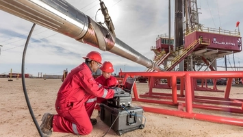 Two workers in red coveralls operating a portable equipment case beside a large drill pipe at an active rig